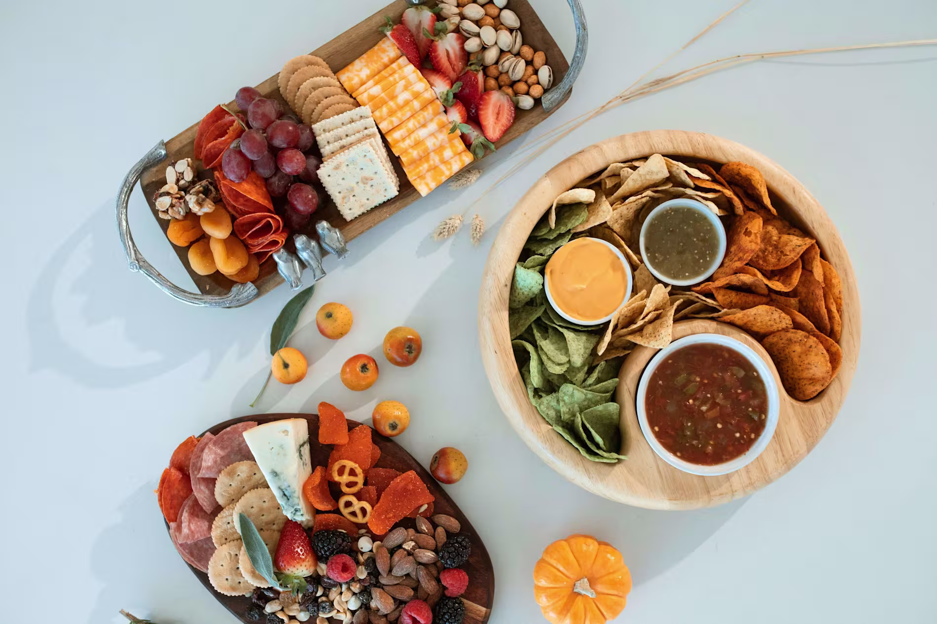 A tabletop with two charcuterie boards featuring meats, cheeses, fruits, and nuts, and a round platter of chips with nacho cheese, salsa, and green dip. Small fruits and a mini pumpkin are scattered around.