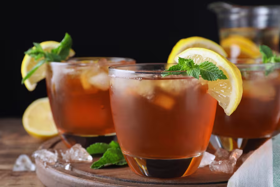 Three glasses of iced tea with lemon slices and mint leaves, served on a wooden tray with ice cubes and fresh mint scattered around.