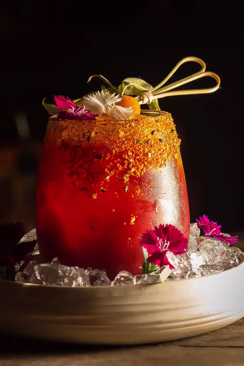 Un cóctel vibrante con tonos rojizos, adornado con flores comestibles, una decoración de frutas, presentado con una base de hielo en CDLC Barcelona.