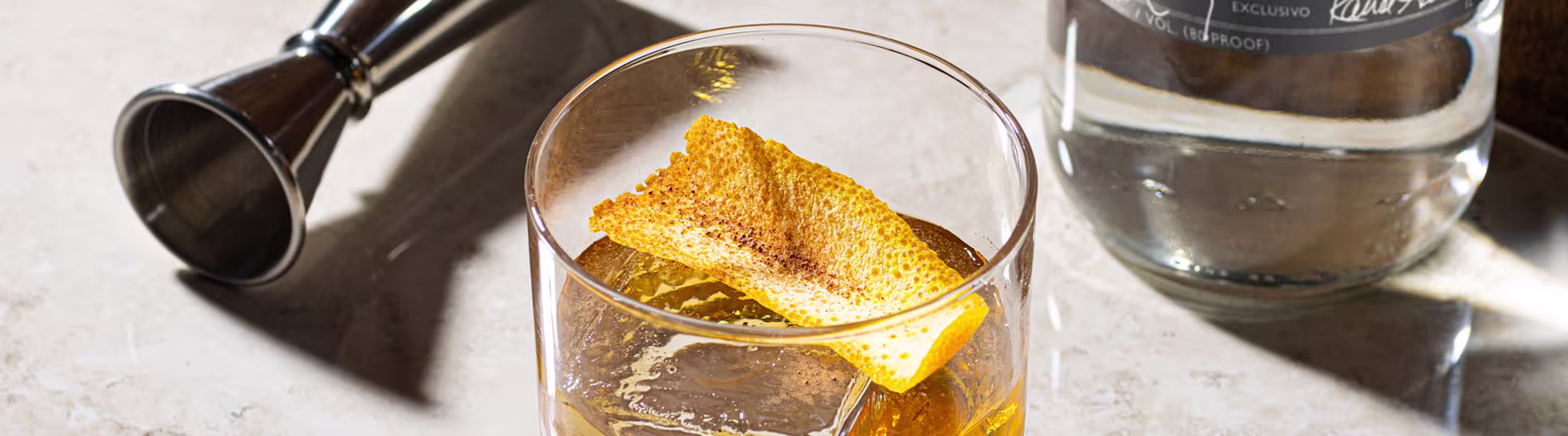 Cóctel a base de agave con guarnición de piel de cítricos servido junto a un jigger y una botella de tequila