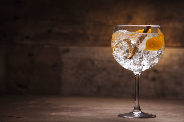 A cocktail glass filled with ice, garnished with orange peel twists and a cinnamon stick, set against a dark wooden background.