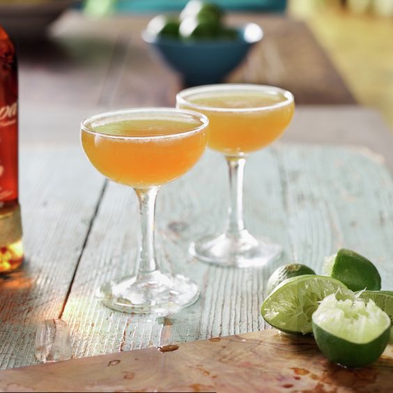 La imagen muestra dos copas rellenas con el cóctel Daiquiri, a su lado, una botella de ron Zacapa 23 y limas exprimidas.