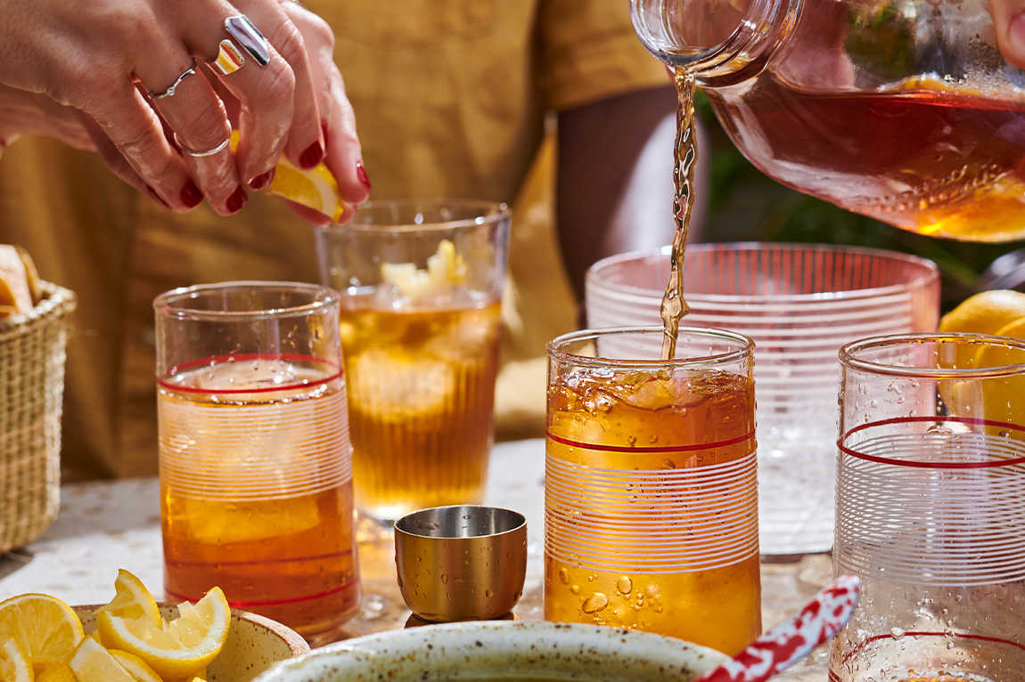 A person pours iced tea into striped glasses while another adds a lemon wedge; sliced lemons and a metal jigger are on the table.