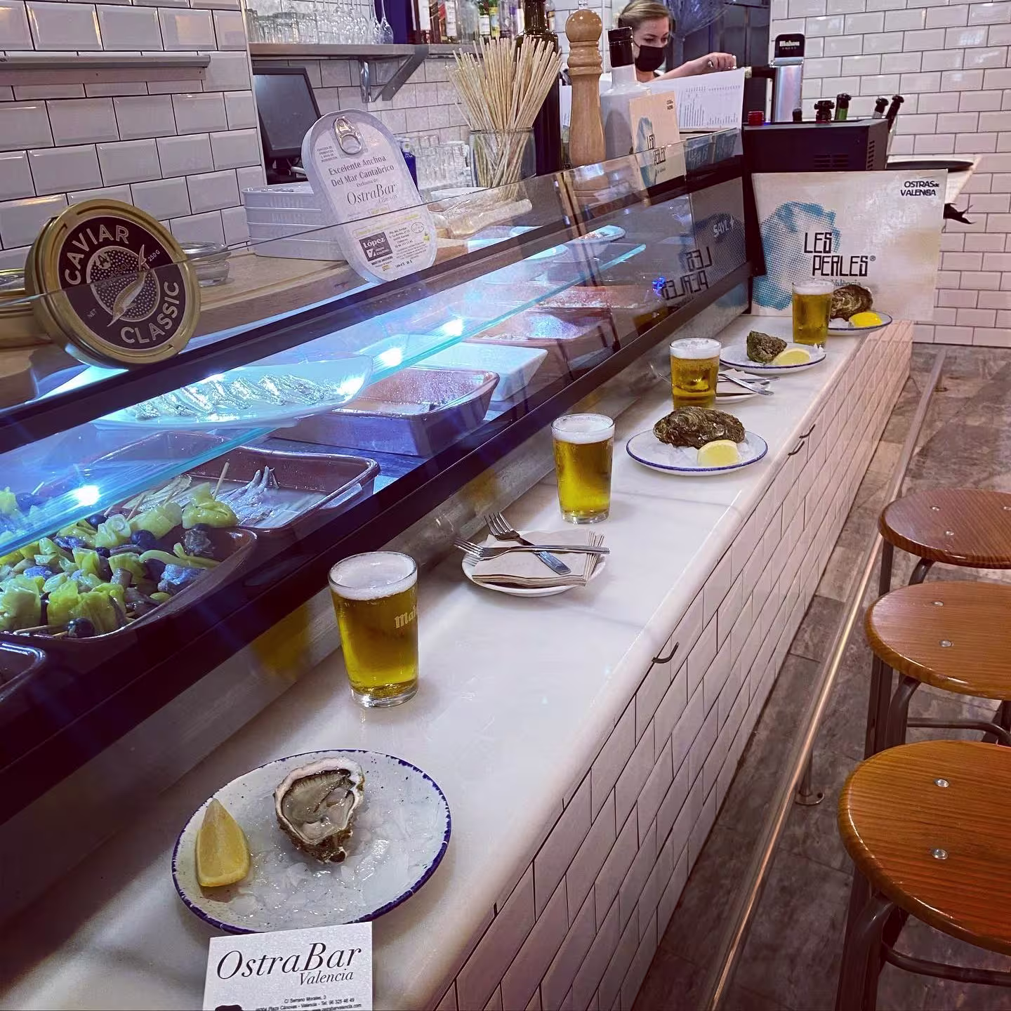 Barra con platos de ostras sobre hielo, cervezas servidas y taburetes de madera. Al fondo, vitrina refrigerada y personal tras la barra.