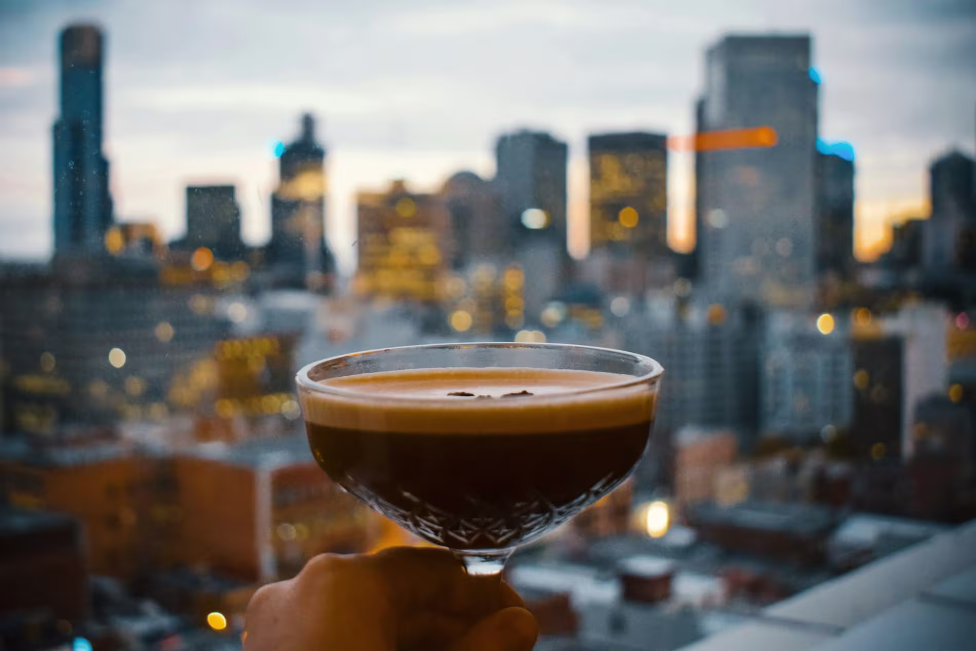 A hand holds a cocktail glass with a dark drink in front of a city skyline at dusk, seen through a window.
