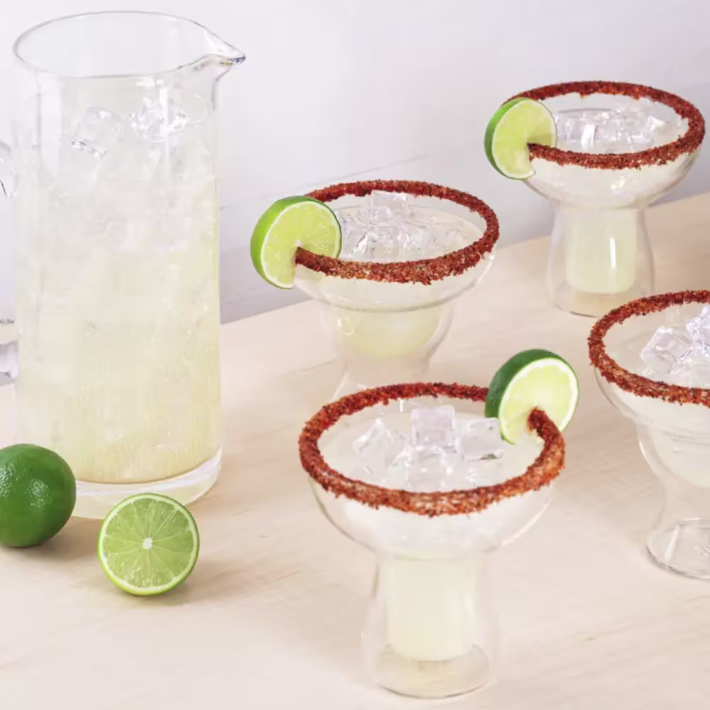 Una jarra y cuatro vasos de margarita con hielo, bordes de chile en polvo y gajos de lima se disponen sobre una superficie clara con dos limas enteras y una mitad de lima cerca.