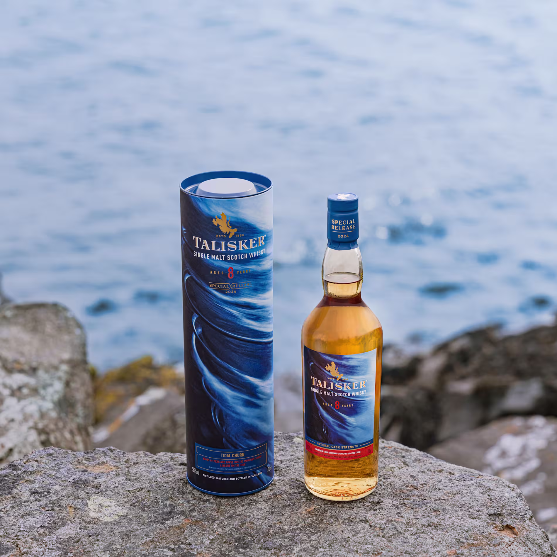 A bottle of Talisker 8-Year-Old Single Malt Scotch Whisky and its blue packaging are placed on a rock near the water’s edge with stones and the sea in the background.