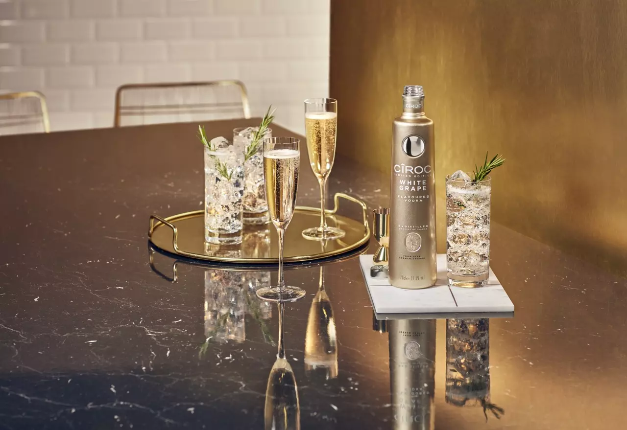 A gold bottle of Ciroc White Grape vodka is placed on a marble table next to two cocktails with ice and rosemary garnish and two champagne flutes, all arranged on trays.