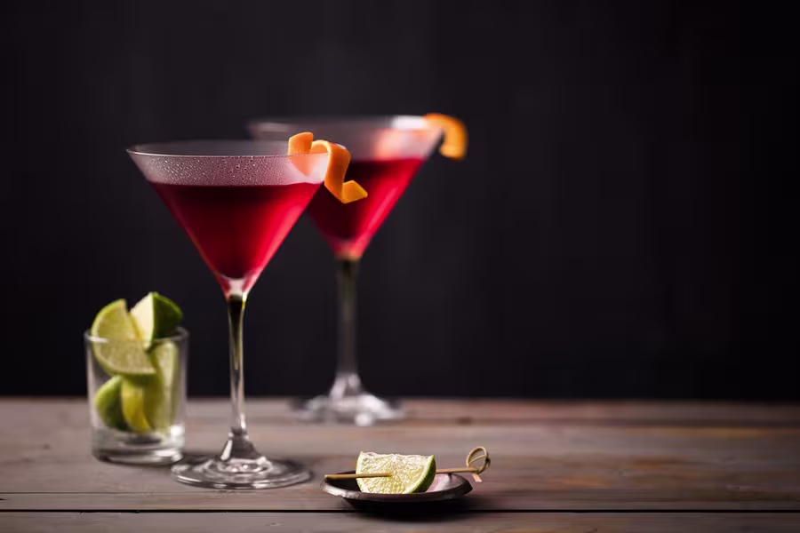 Two red cocktails in martini glasses with orange peel garnishes, next to a glass of lime wedges and a small plate with lime slices, on a wooden surface against a dark background.