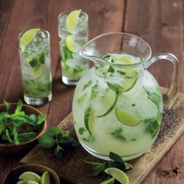 A pitcher filled with a refreshing cocktail garnished with lime slices and fresh mint leaves, accompanied by two tall glasses containing the same cocktail. 
