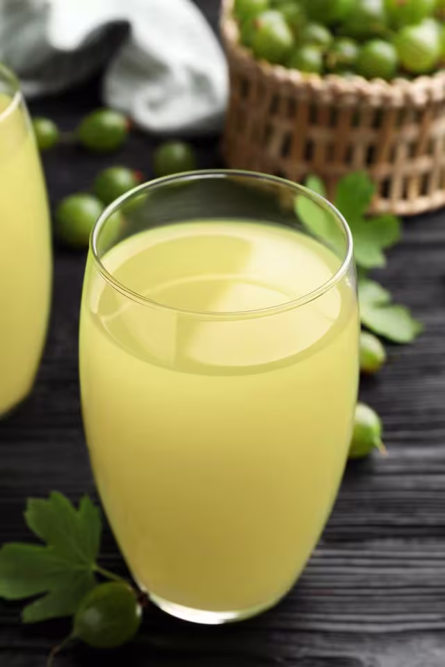 A glass of pale yellow gooseberry juice on a dark surface, with fresh gooseberries and a basket in the background.