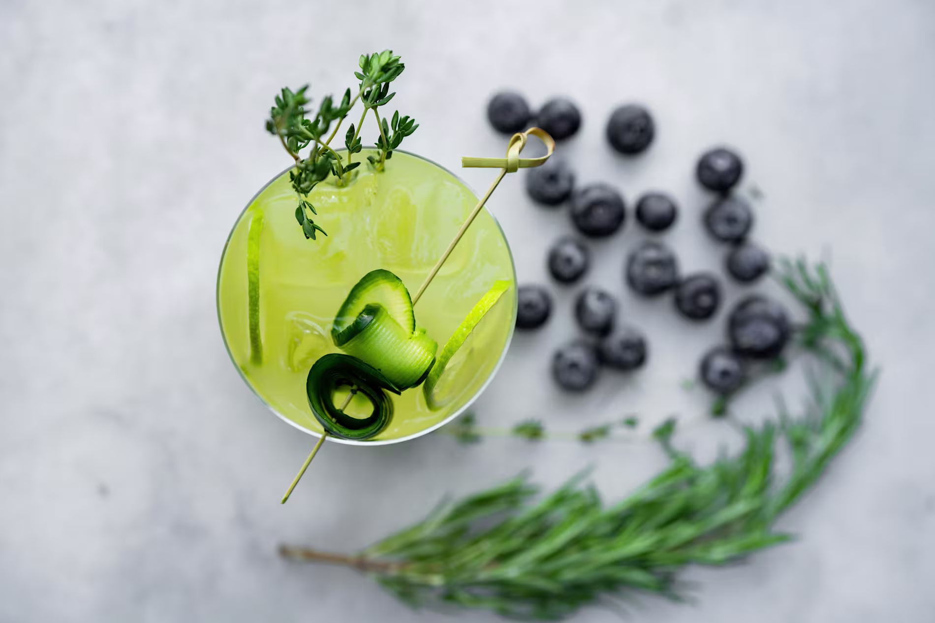 A green cocktail with ice, cucumber ribbons, and a sprig of herbs, viewed from above, with blueberries and a rosemary sprig on a light surface nearby.