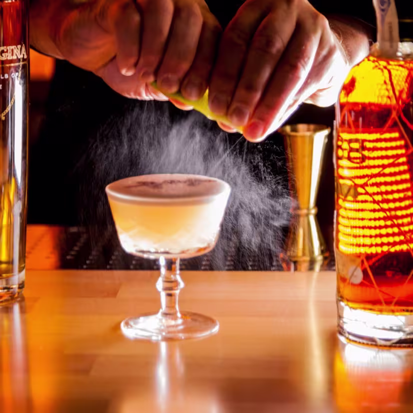 Un cóctel elegante en plena creación, con luz cálida y detalle artesanal en cada gesto del bartender.