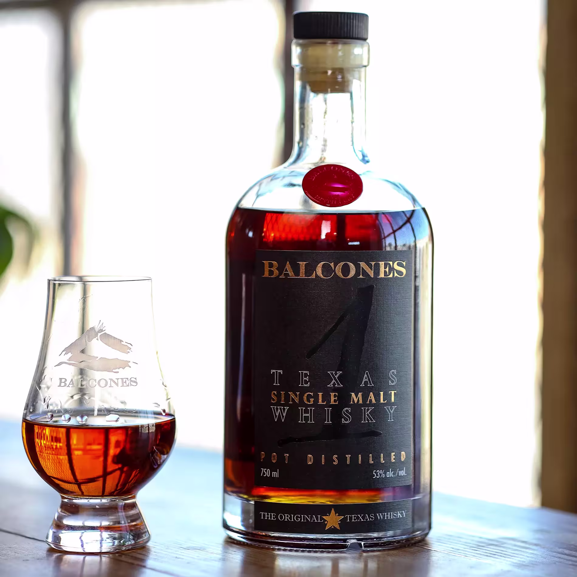 A bottle of Balcones Texas Single Malt Whisky and a filled whisky glass sit on a wooden surface in sunlight.