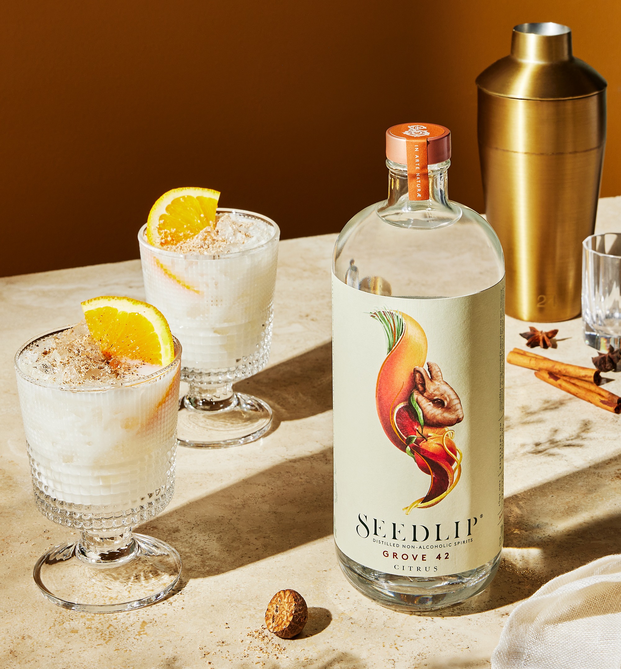 A bottle of Seedlip Grove 42 stands beside two glasses of creamy white cocktails garnished with orange slices and grated spice, with a cocktail shaker and star anise in the background.