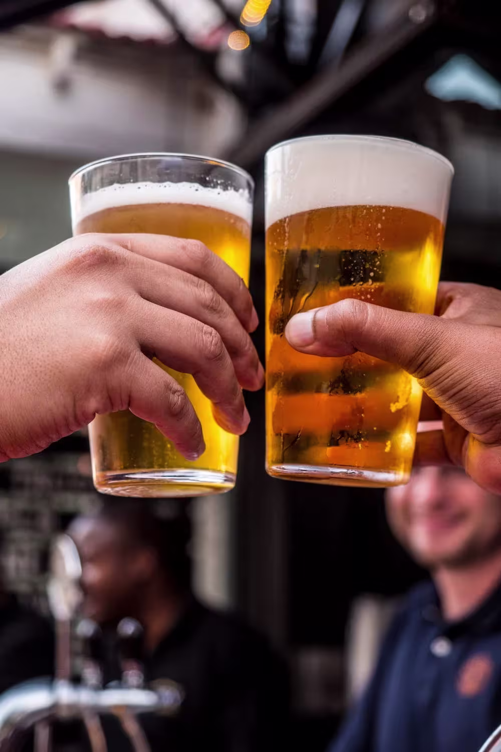 Two people clink glasses of beer together, with foam visible at the top, in an outdoor setting with blurred people in the background.