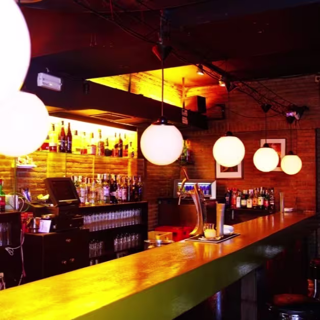 La imagen muestra el interior de Honky Tonk. La barra está iluminada por luces superiores, y hay varias botellas de alcohol exhibidas en estantes contra una pared de ladrillo. Grandes luces esféricas blancas cuelgan del techo, añadiendo al ambiente. 