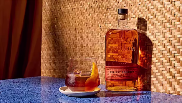 A bottle of Bulleit Bourbon whiskey stands next to a glass of whiskey with ice and an orange twist, placed on a blue surface against a woven background.