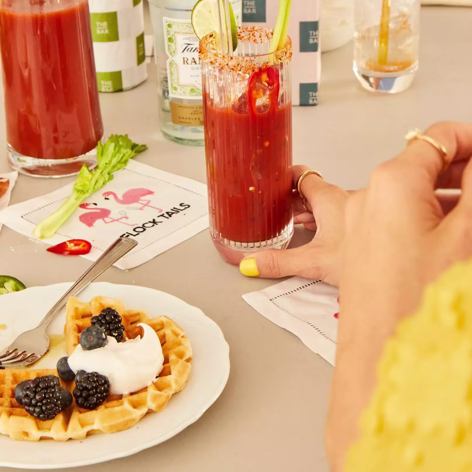 A hand reaches for a Bloody Mary cocktail garnished with lime and chili, next to a plate with a waffle topped with whipped cream and berries.