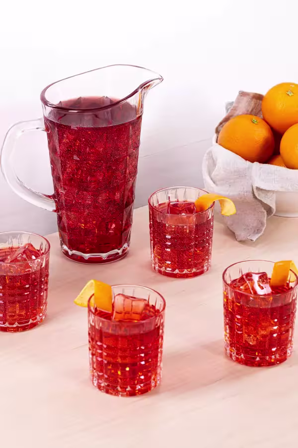 A pitcher and four glasses of red cocktail with orange peel garnish are on a light surface next to a white bowl of oranges.
