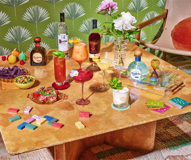 A table set with various cocktails, liquor bottles, snacks, a vase of flowers, dominos, and playing cards, against a green patterned wallpaper background.