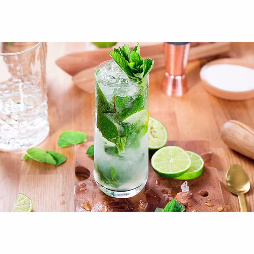 En la foto se aprecia un vaso con el cóctel Mojito, y en el fondo, hay elementos de coctelería relacionados con este trago.