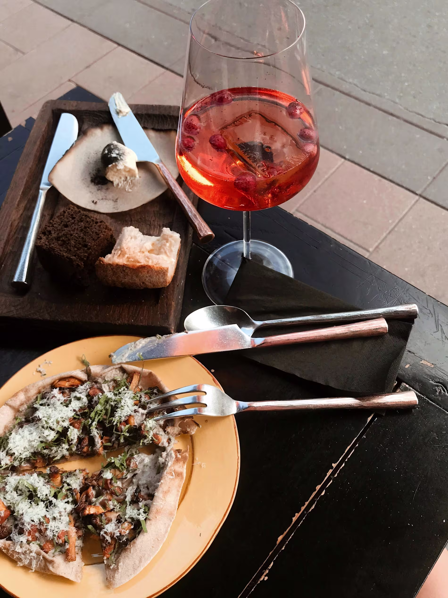 A plate with sliced flatbread topped with herbs and cheese, a wooden board with bread and butter, and a glass of orange cocktail with berries on a black table.
