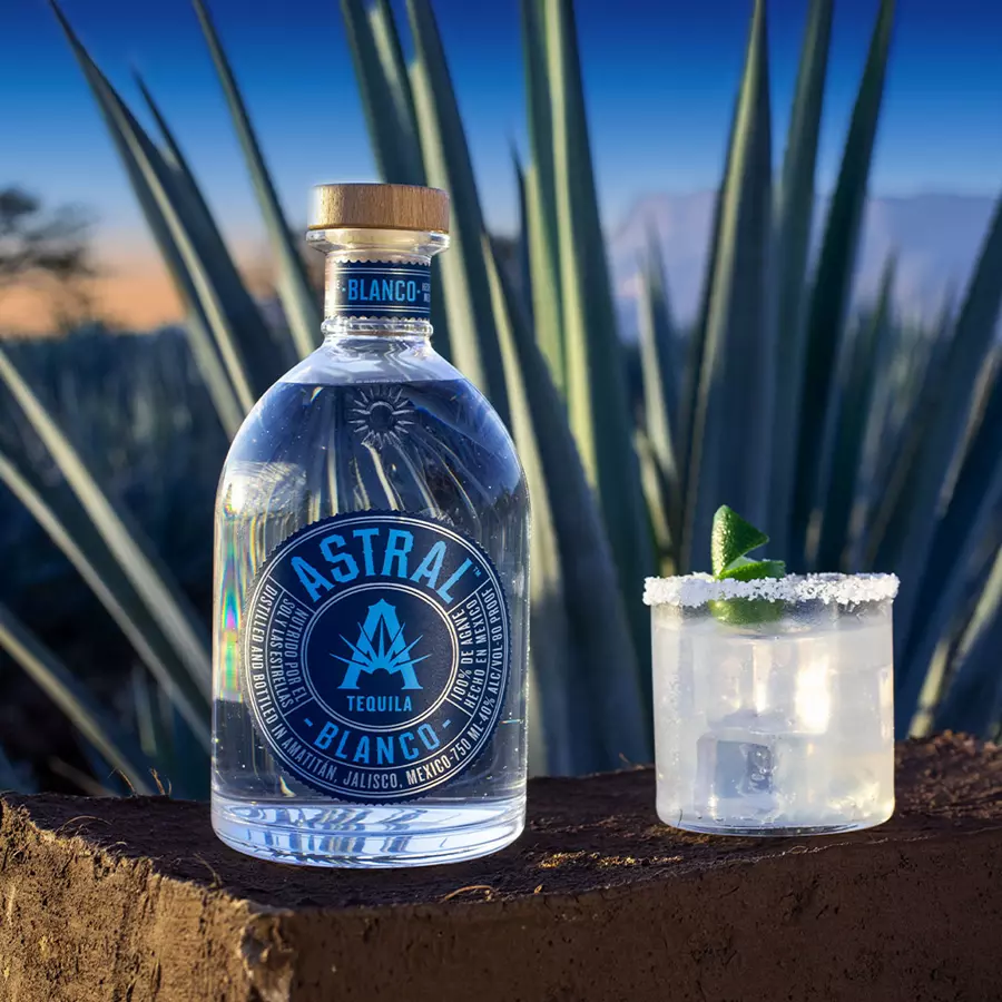 A bottle of Astral Blanco Tequila and a salt-rimmed glass with a lime garnish sit on a brick ledge in front of agave plants.