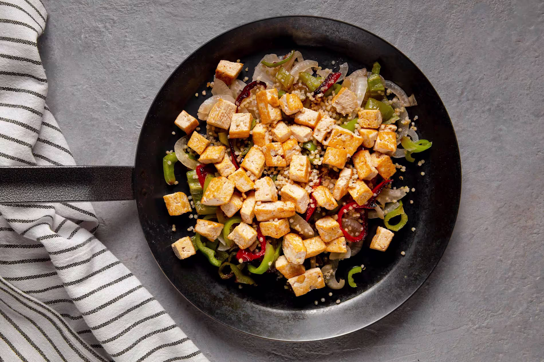 A black skillet with cooked cubed tofu, onions, green peppers, and sliced red chili peppers, placed on a gray surface next to a striped kitchen towel.