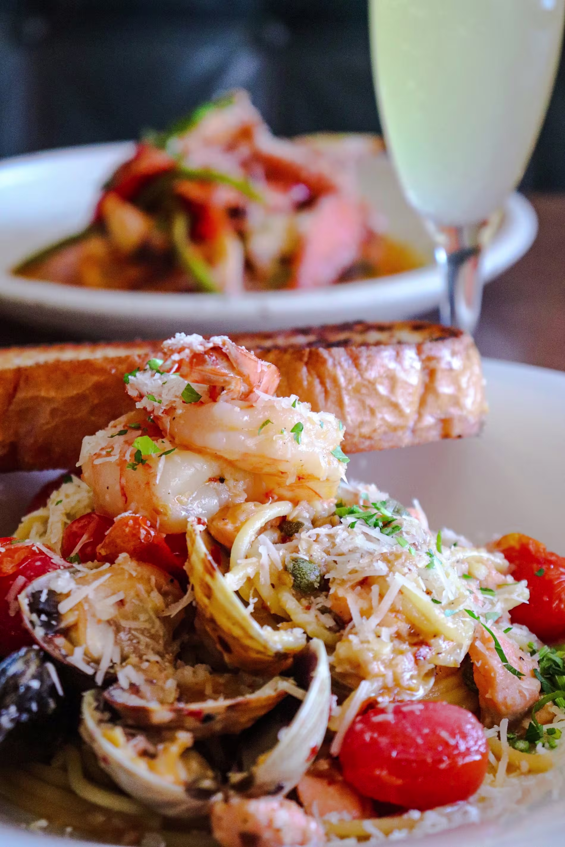 A plate of seafood pasta with shrimp, clams, cherry tomatoes, and grated cheese, served with toasted bread; a drink and another dish are in the background.