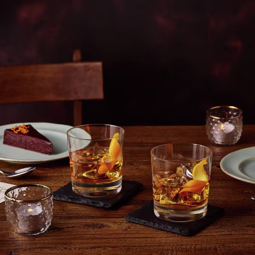 La imagen muestra dos vasos con el cóctel Old Fashioned, así como dos platos con una porción de torta cada uno y dos cucharas.