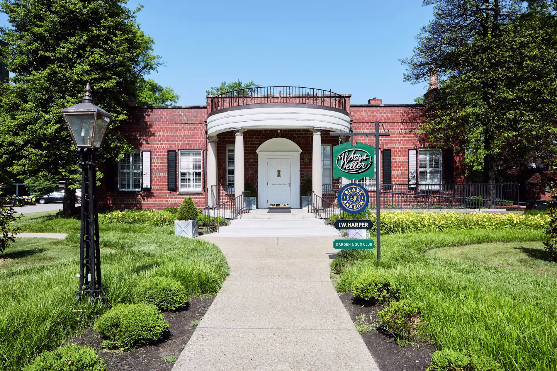 Single-story red brick building with white columns and a white door, surrounded by trimmed bushes; a real estate sign is visible near the entrance walkway.