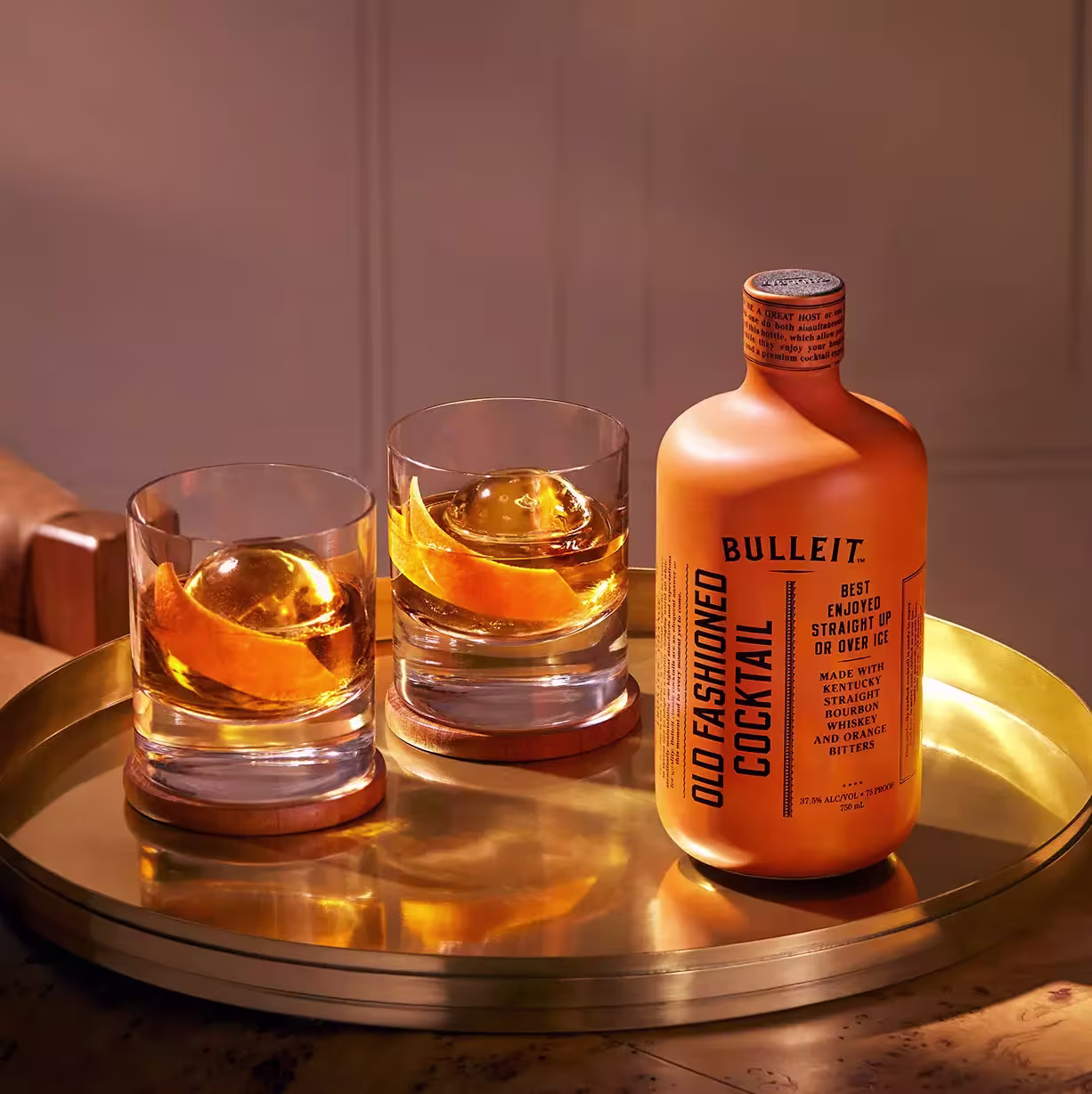 A gold tray holds a bottle of Bulleit Old Fashioned Cocktail and two glasses filled with whiskey, large ice spheres, and orange peels.