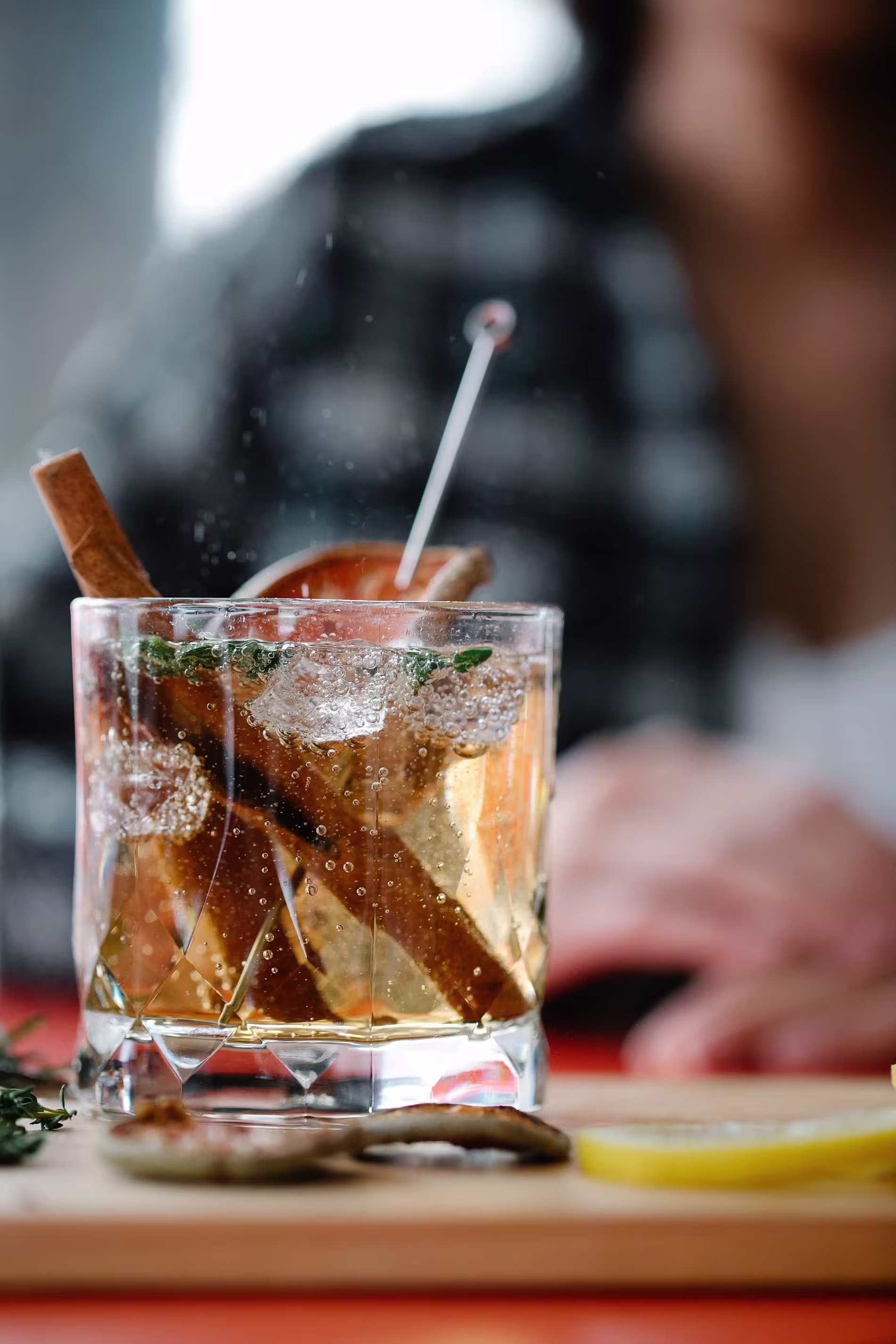 A glass of iced cocktail garnished with cinnamon sticks, herbs, and an orange slice sits on a table near lemon wedges, with a blurred person in the background.