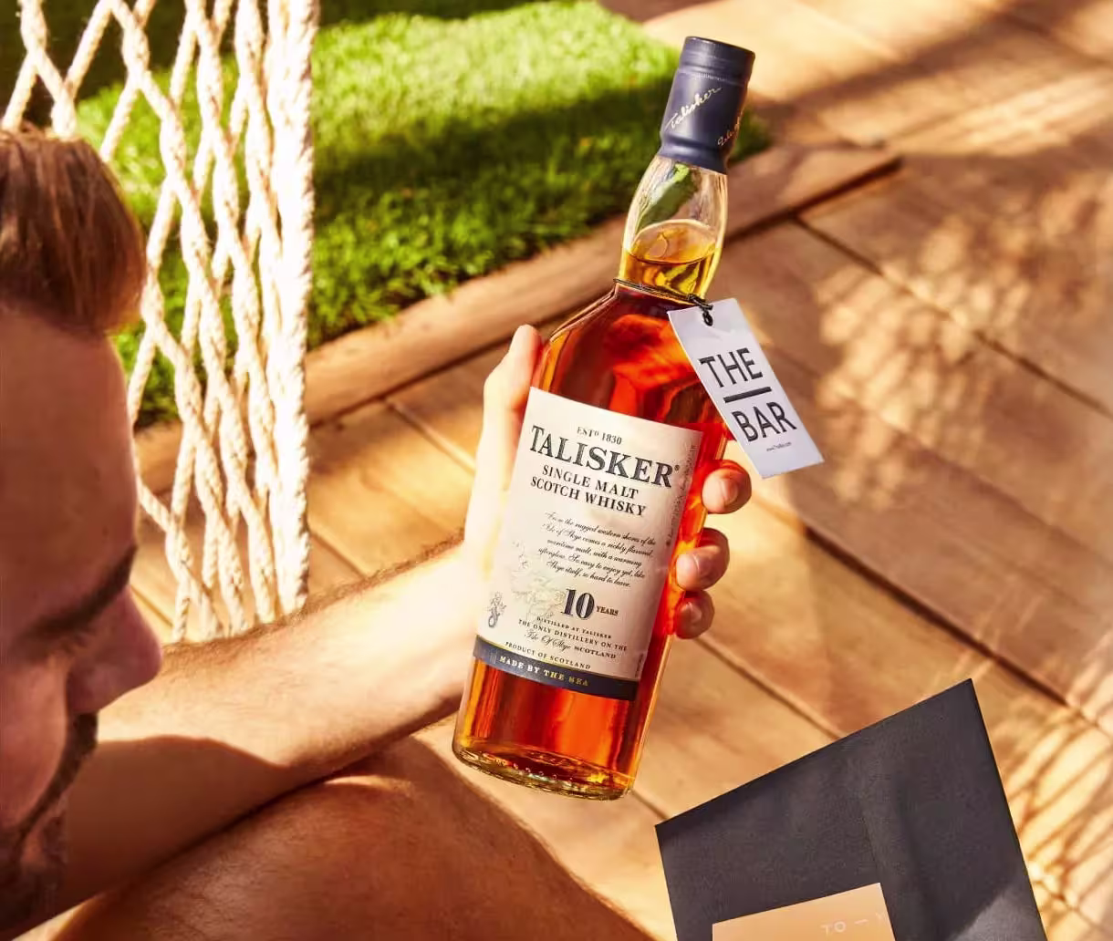 A person holding a bottle of Talisker 10 Year Old Single Malt Scotch Whisky while sitting on a wooden deck.