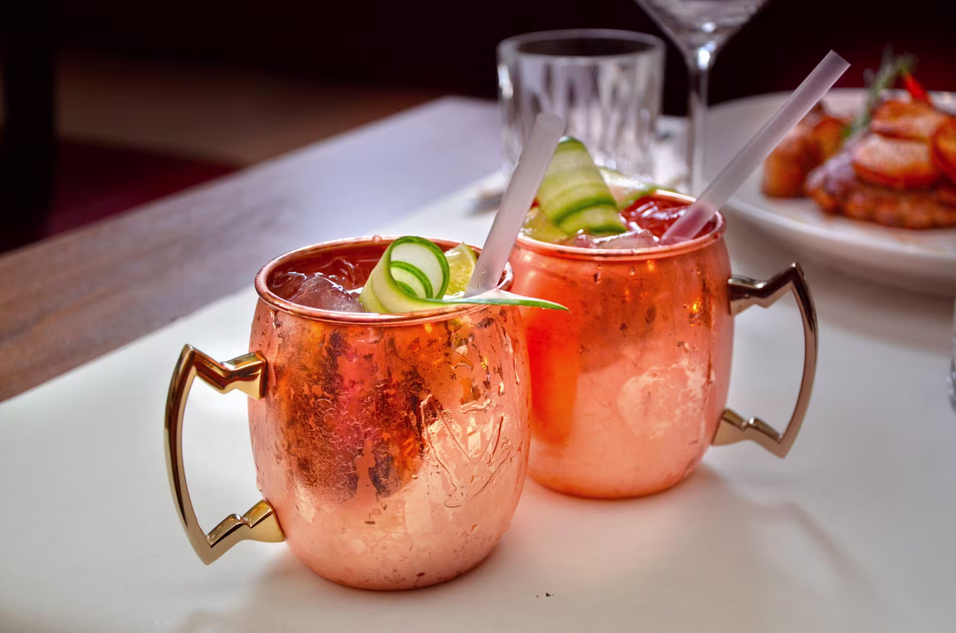 Two copper mugs filled with iced cocktails, garnished with cucumber and lime slices, sit on a white table next to glassware and a plate of food in the background.