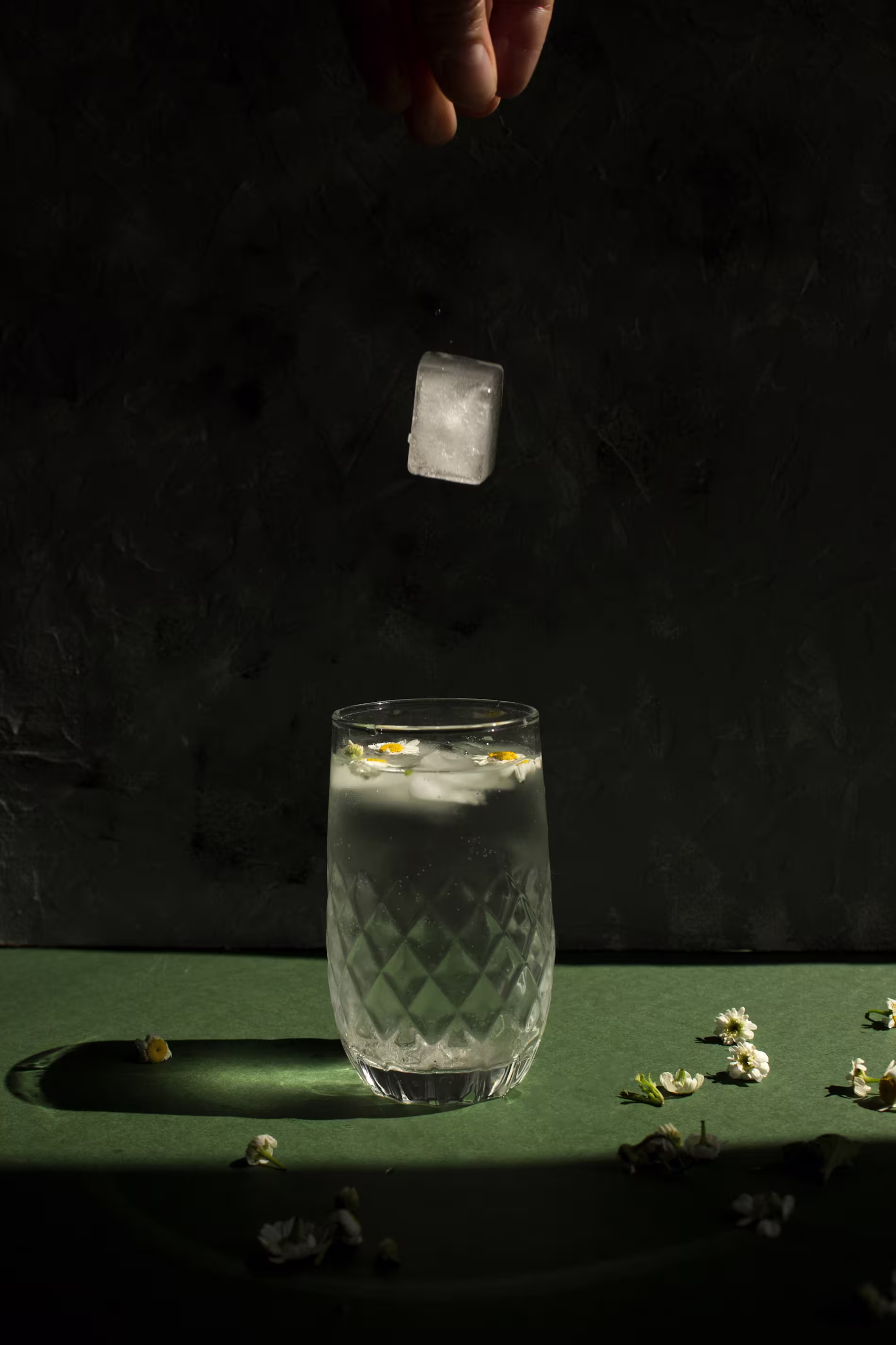 A hand drops an ice cube into a clear, patterned glass of water with flowers floating on top, set on a green surface with scattered flower petals.