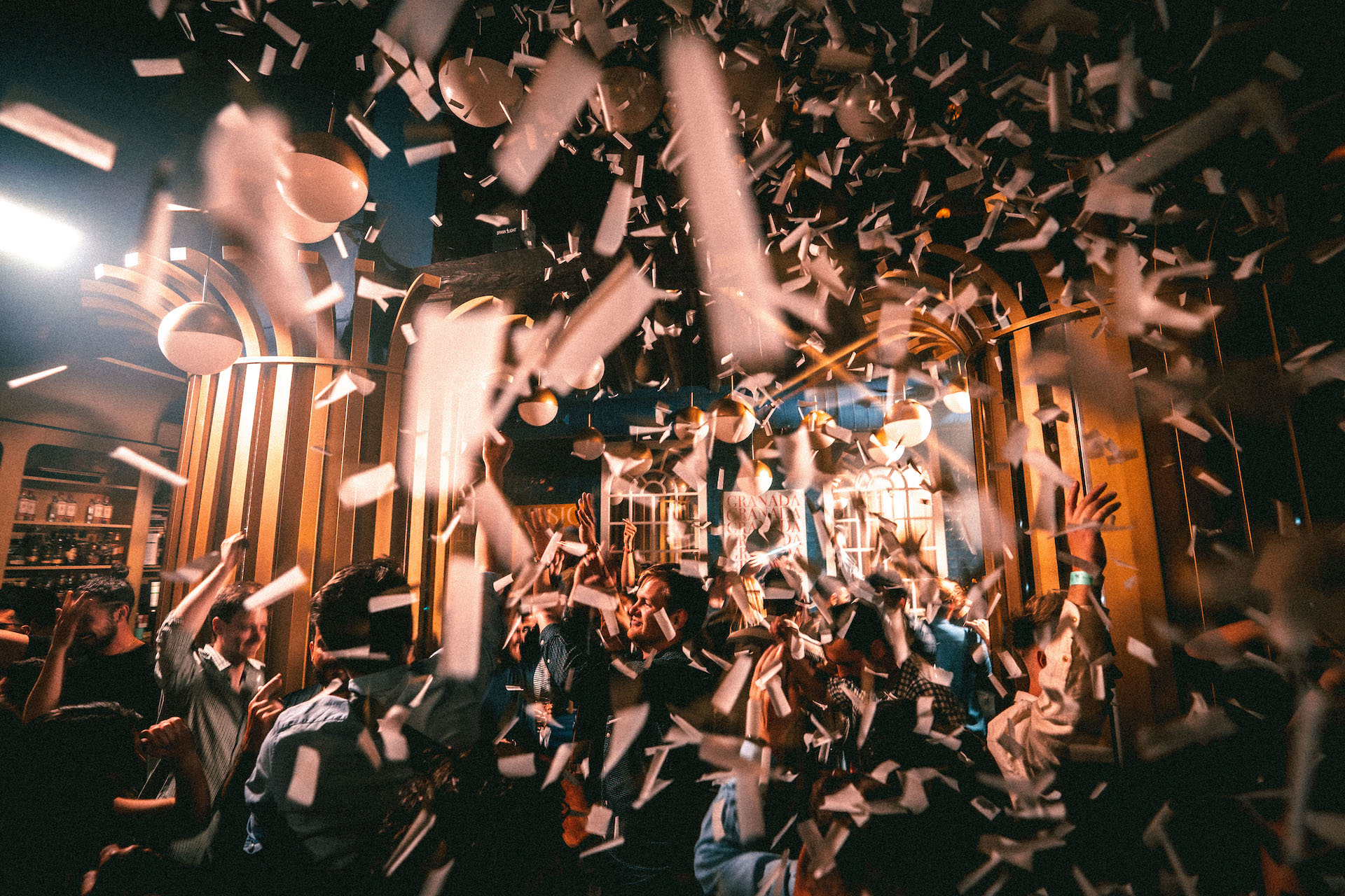La imagen captura una fiesta vibrante con confeti cayendo sobre una multitud animada. Las luces cálidas y la decoración dorada del lugar crean un ambiente elegante y festivo, lleno de energía y movimiento.