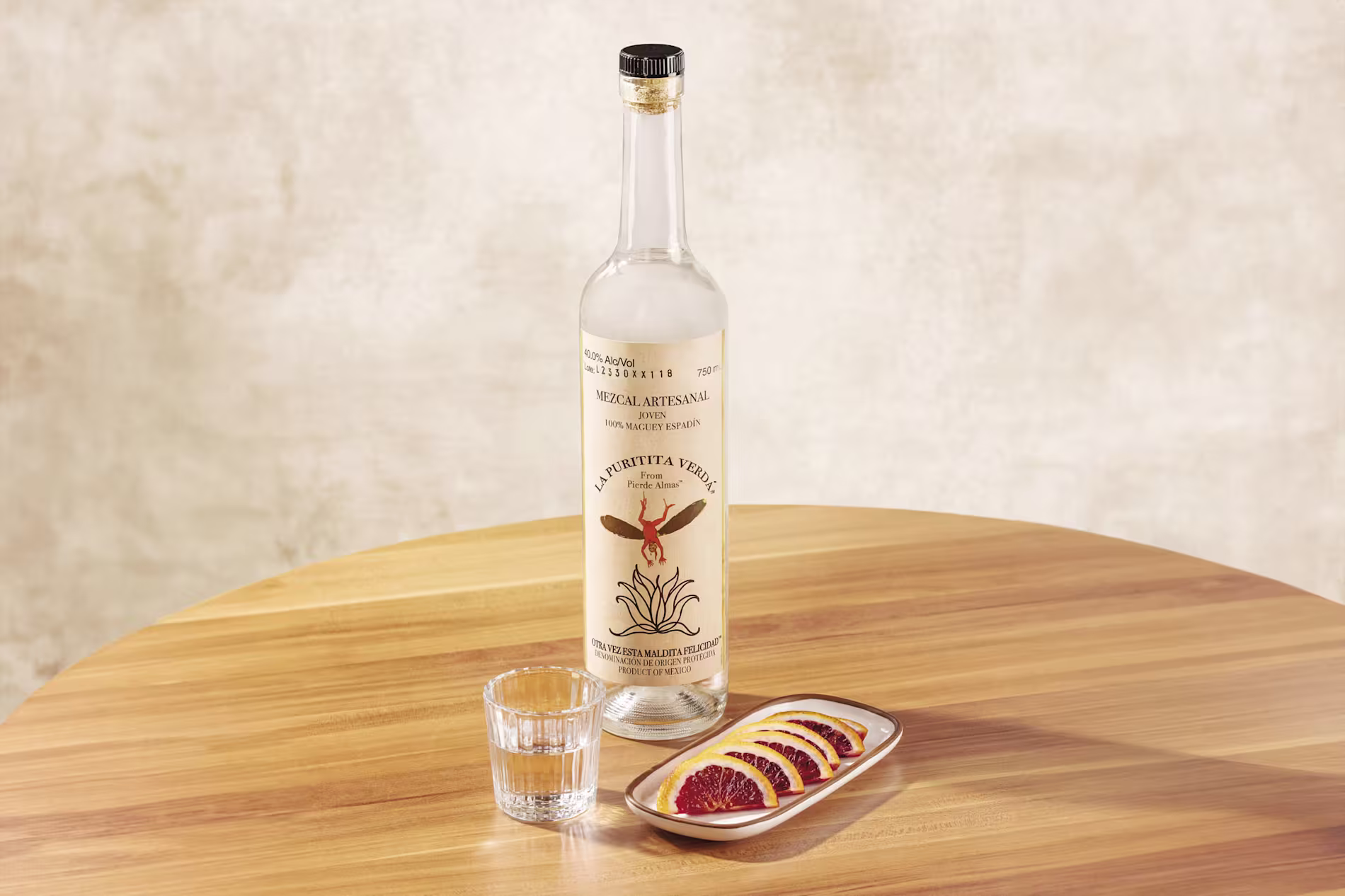 A bottle of mezcal, a clear glass of liquor, and a plate of orange slices are arranged on a wooden table against a neutral background.