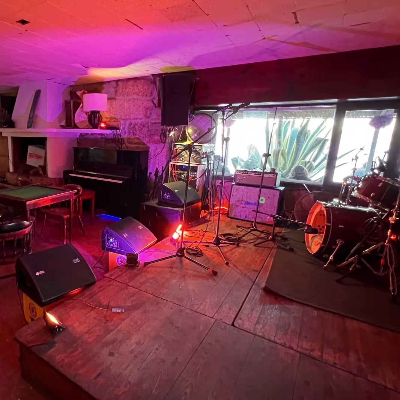 Escenario en Náutico San Vicente con micrófonos, batería y amplificadores listos para una actuación. Ambiente acogedor con muebles rústicos y un piano en la esquina. Ventana amplia al fondo aporta luz natural."