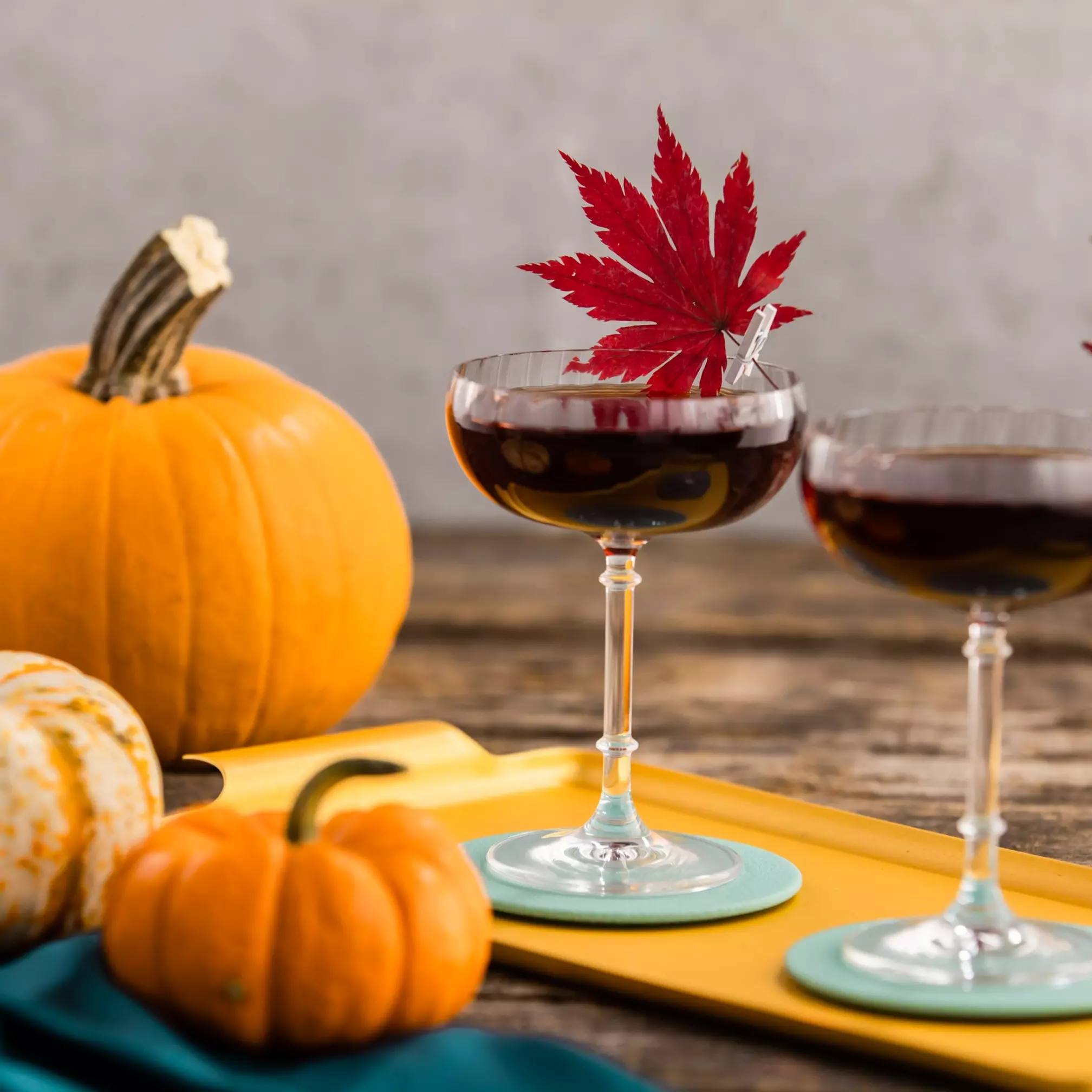 Two cocktail glasses with dark liquid and red maple leaf garnishes sit on a yellow tray next to three pumpkins on a wooden surface.