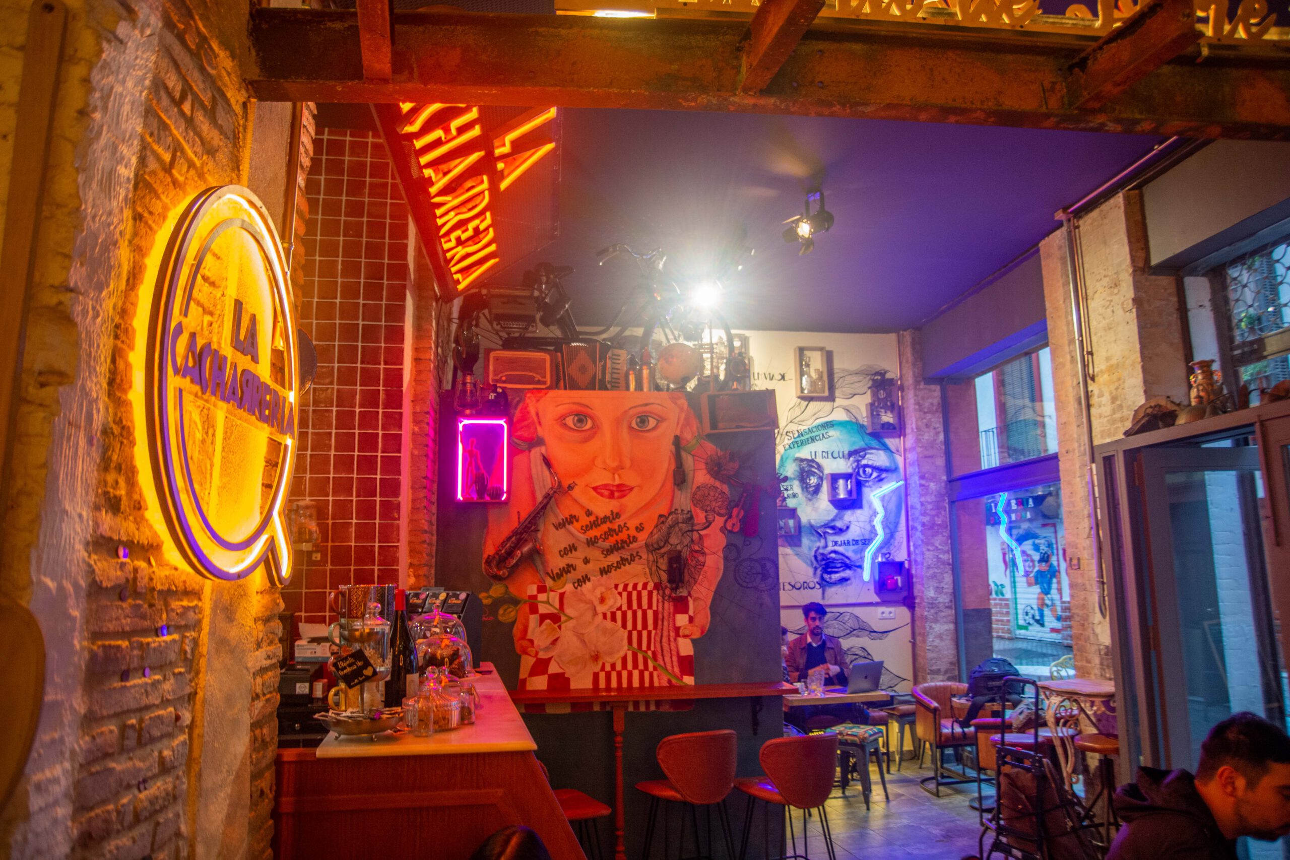 Interior de La Cacharrería con murales coloridos, luces de neón, mobiliario ecléctico y barra con café y botellas sobre una pared de ladrillo.