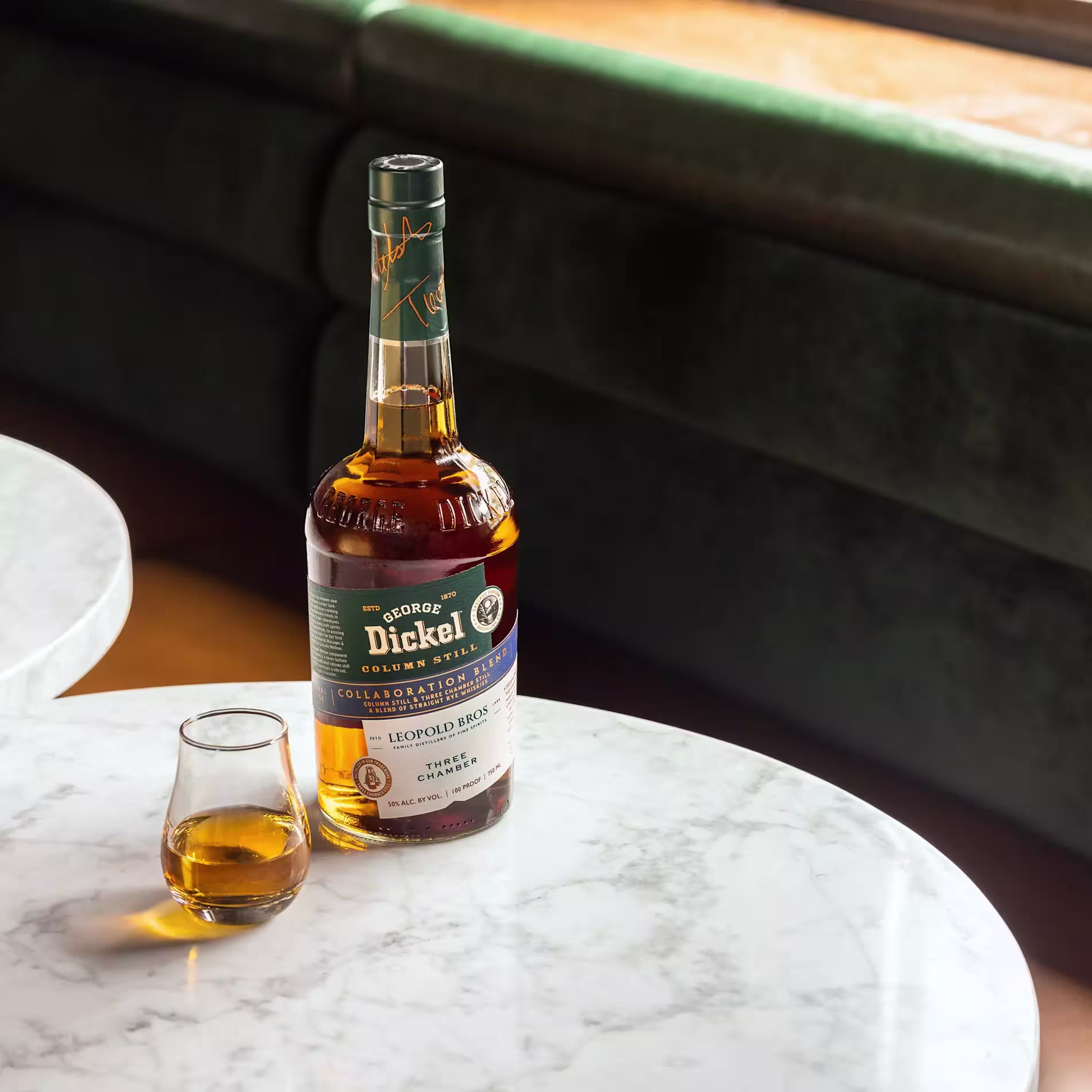 A bottle of George Dickel bourbon whiskey and a glass with whiskey sit on a round white marble table next to a green cushioned bench.