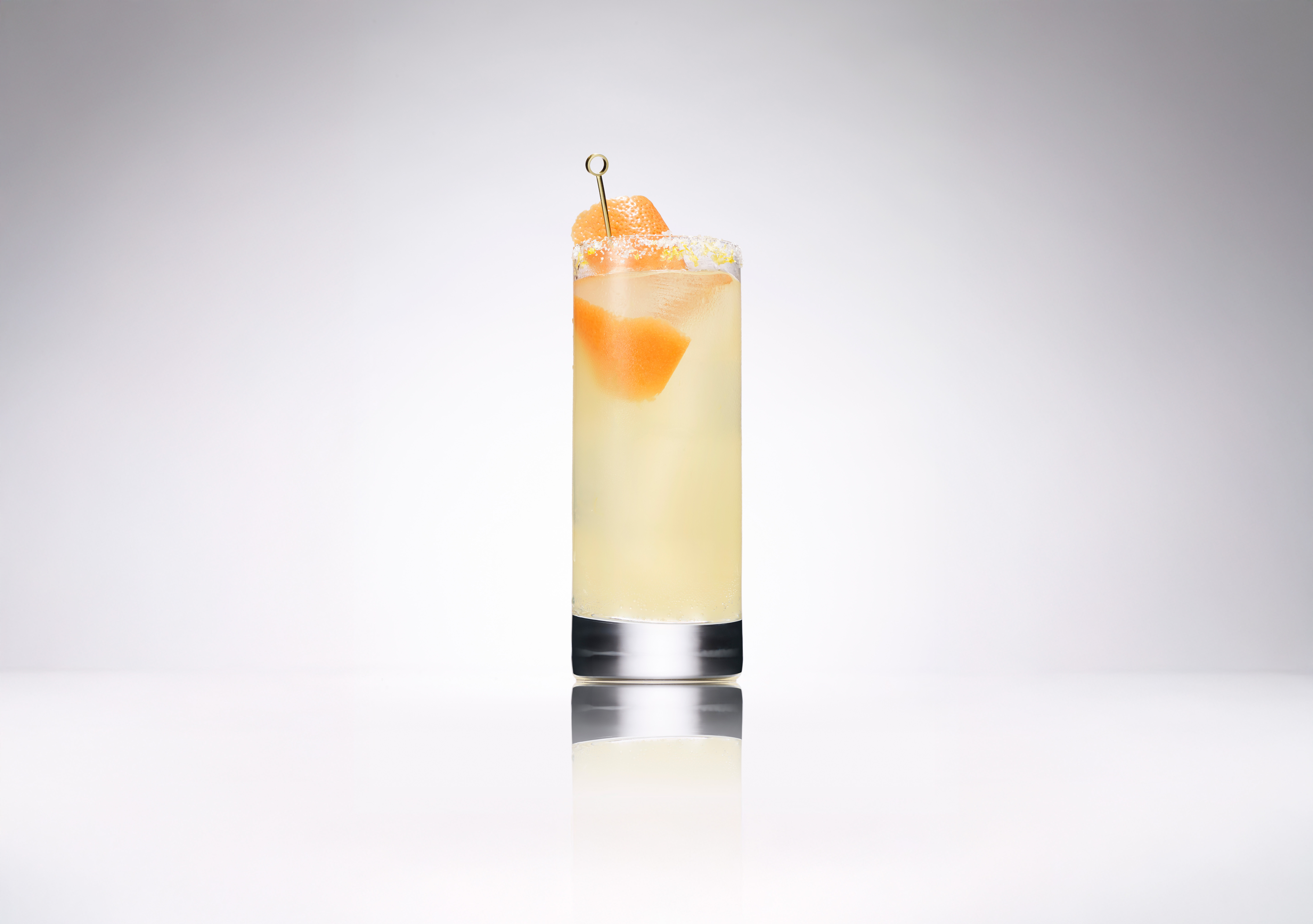 A tall glass of pale yellow cocktail garnished with two grapefruit wedges on a skewer, set against a plain white background.