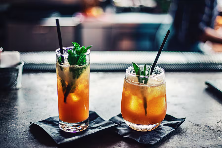 Two orange cocktails with black straws and green garnish sit on black napkins on a bar counter, with a blurred background.