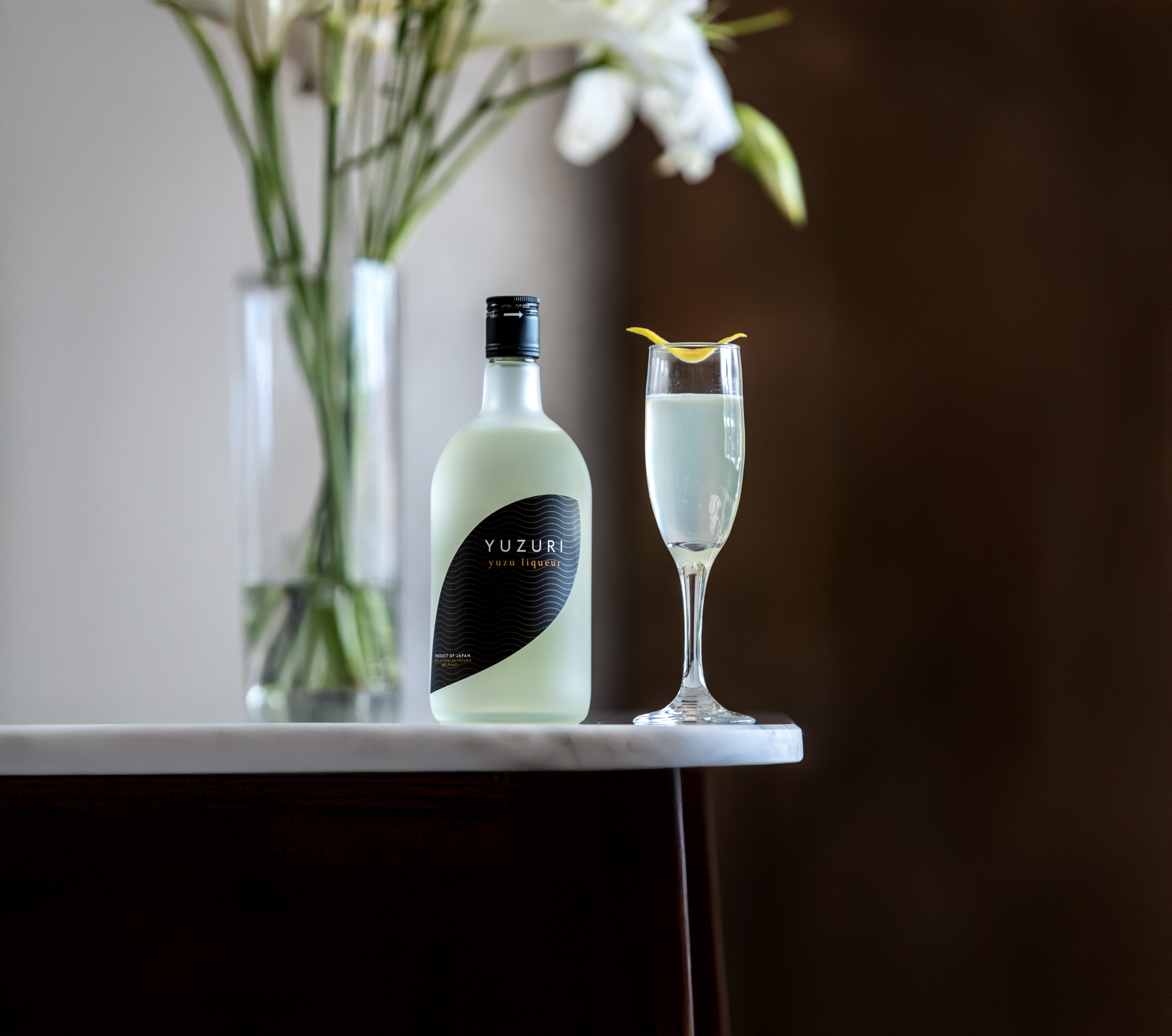 A bottle of Yuzuru Yuzu Liqueur and a cocktail garnished with a lemon twist are placed on a marble surface next to a vase of white flowers.