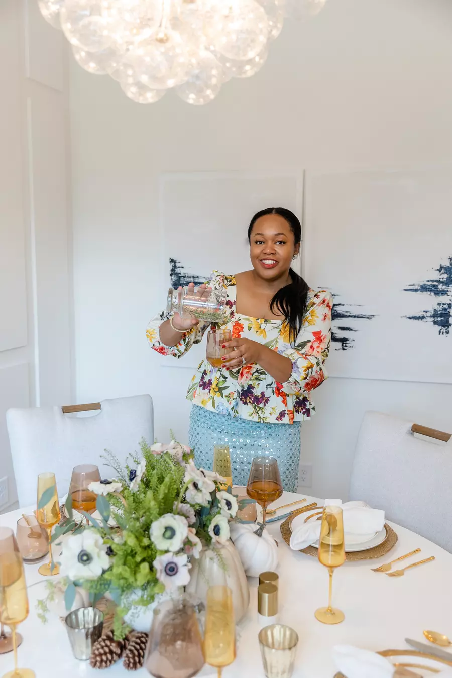 A woman in a floral blouse pours a drink at a decorated dining table set with flowers, glassware, and gold utensils under a modern chandelier.