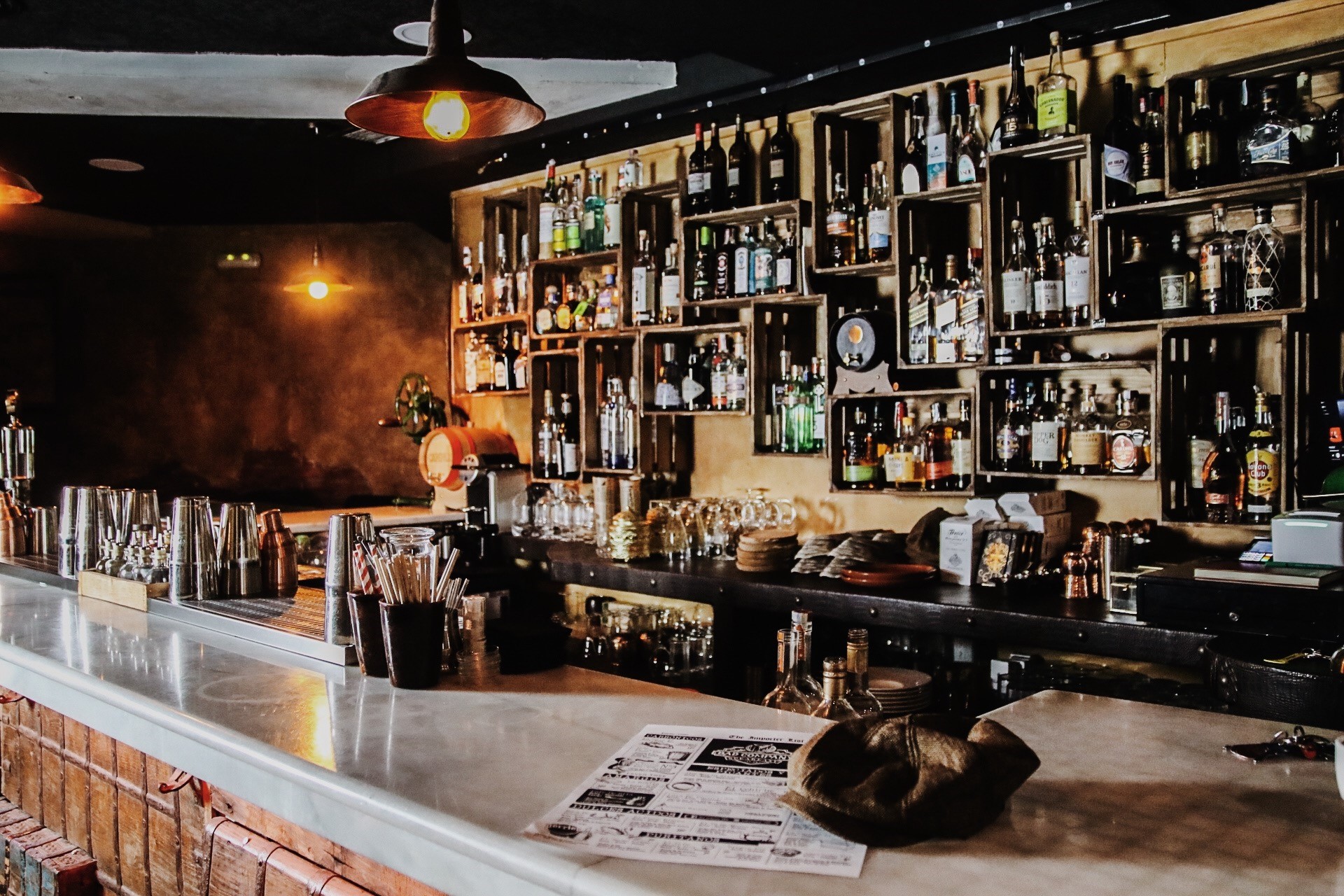 El interior del "Bad Company 1920" con una variedad de botellas en estantes contra una pared oscura, iluminada con una luz cálida. En primer plano hay una encimera blanca con varios artículos como pajitas y servilletas organizados.