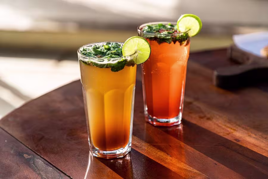 Two tall glasses of iced drinks, one orange and one red, garnished with lime slices and mint, sit on a wooden table in sunlight.