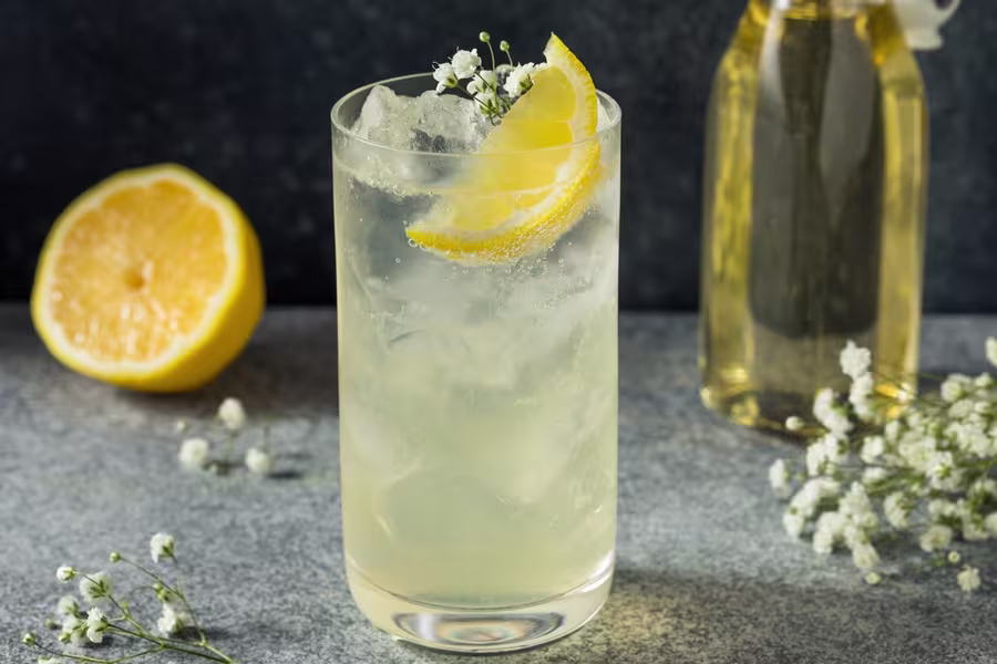A glass of iced lemon drink garnished with a lemon slice and small white flowers, with a halved lemon and a glass bottle in the background.